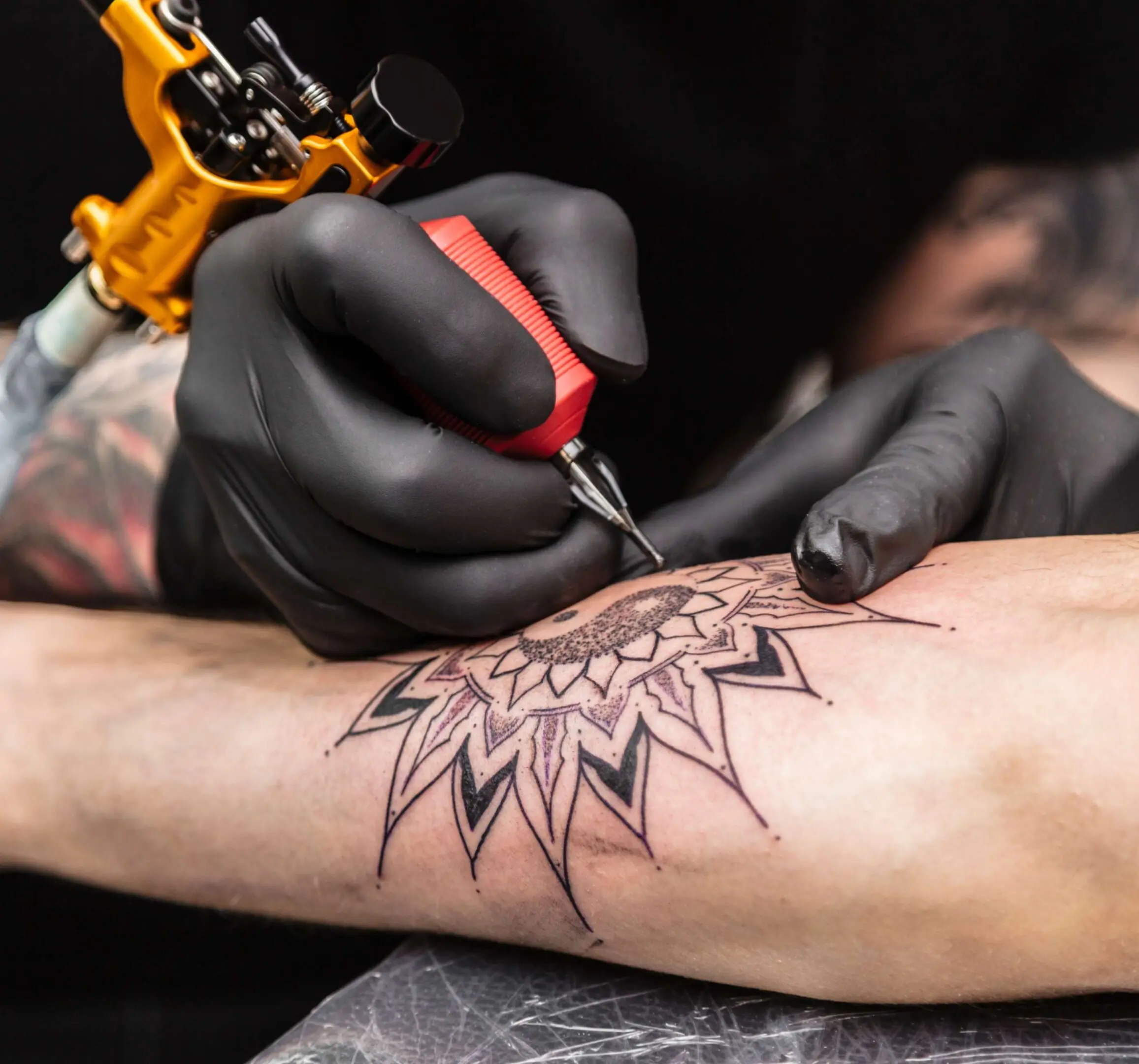 jpeg-optimizer_GettyImages-1162623180 Tattoo artist with black gloves is applying a symmetrical, detailed sun-like design on a person's forearm using a tattoo machine.
