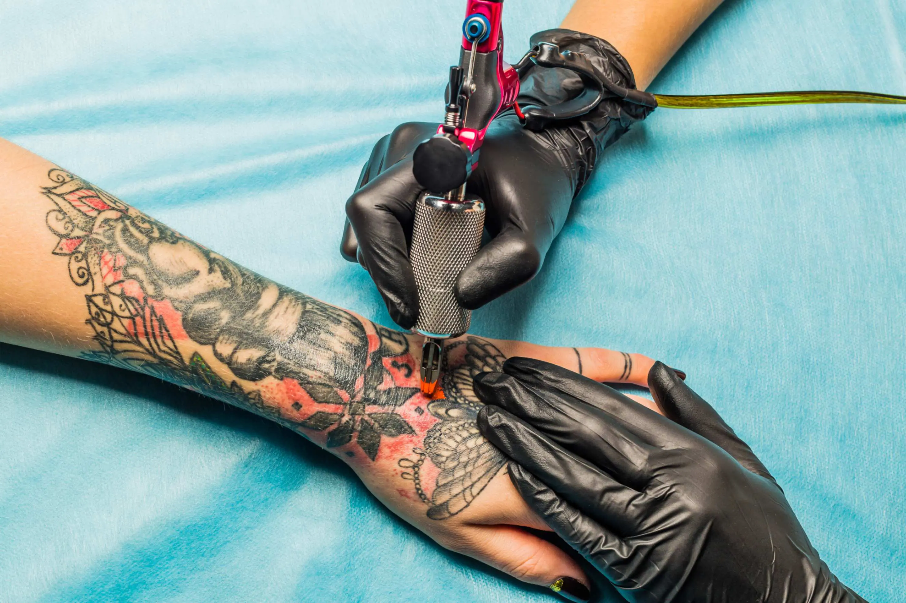 jpeg-optimizer_GettyImages-491837154 A tattoo artist wearing black gloves uses a tattoo machine to ink a colorful design on a person's forearm.