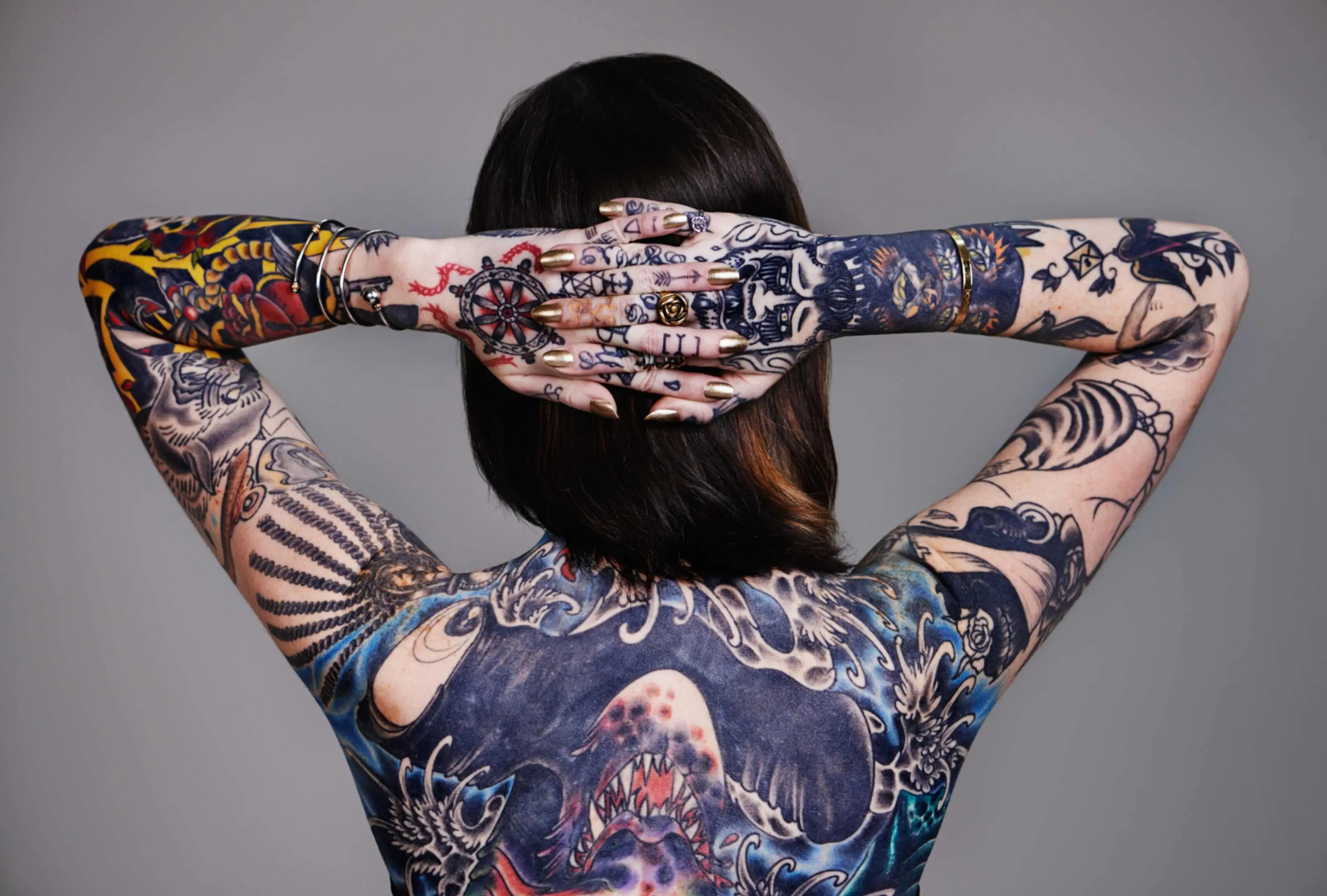 jpeg-optimizer_GettyImages-520866989 Person facing away, arms raised and crossed behind their head, displaying extensive, colorful tattoos covering back, arms, and hands.