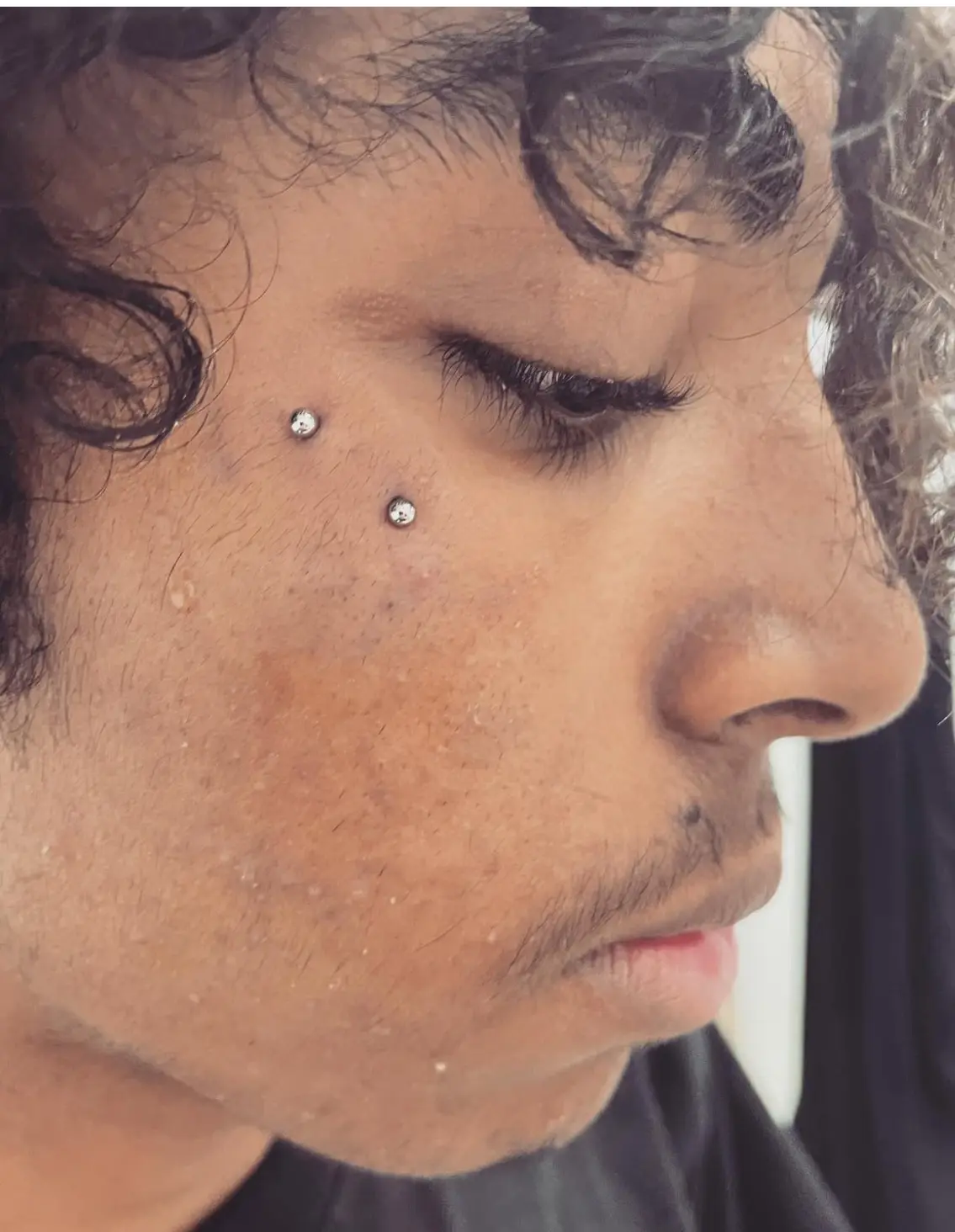A man with curly hair and two piercings on his face.